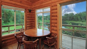 Interior de una cabaña de madera con una mesa redonda y cuatro sillas, ventanas amplias que dan a una zona boscosa.