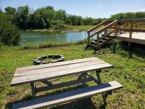 Een houten picknicktafel met een vuurkuil, een houten terras en uitzicht op een meer omringd door bomen.