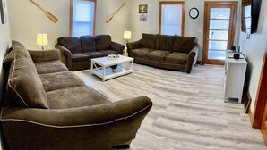 Un salon avec deux canapés marron, une table basse blanche et une petite table d’appoint blanche. Il y a deux fenêtres avec des stores, une horloge au mur et un téléviseur fixé au mur.