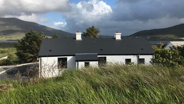 Un edificio blanco con techo oscuro, rodeado de césped y árboles, con montañas al fondo.