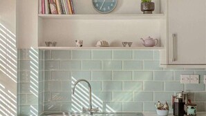 A kitchen with a sink, a clock, and a shelf with books and decorative items. - Apple Cottage - Central Frome - Family Friendly (Somerset)