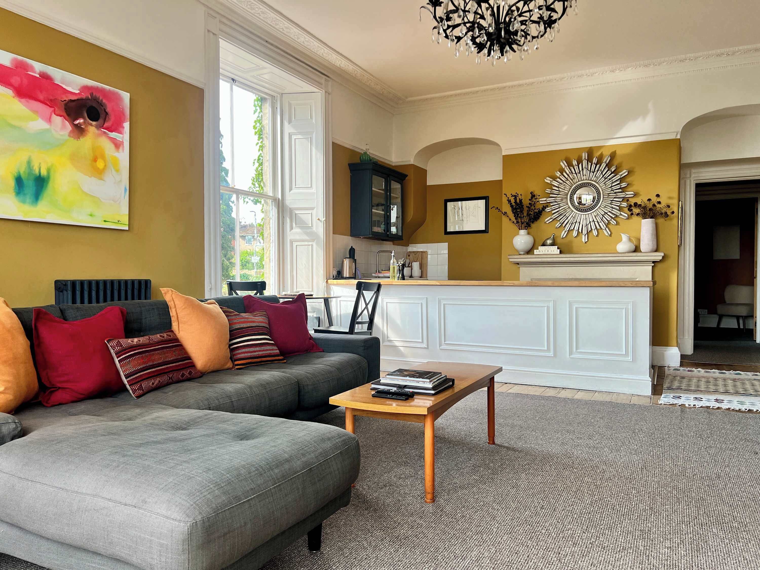 A living room with a grey sofa, a coffee table, and a painting on the wall. There is a kitchen area with a white cabinet and a decorative mirror.