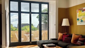 Ein Wohnzimmer mit einer Couch, einem Couchtisch und einer Lampe. Ein großes Fenster bietet Blick auf einen Garten und einen Zaun.