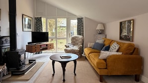A living room with a sofa, a television, a fireplace, and a large window. - The View Cottage -Tennis Court- Nr Frome, Longleat (Nunney)