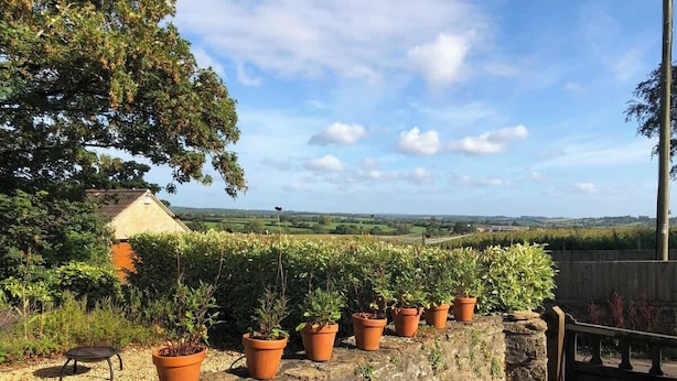 A stone wall with potted plants, a small table, and a house in the background. - The View Cottage -Tennis Court- Nr Frome, Longleat (Nunney)