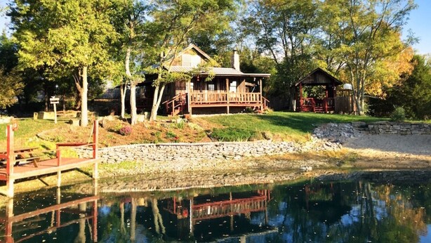 Une cabane rustique avec une terrasse en bois, un mur de pierre et un étang au premier plan.