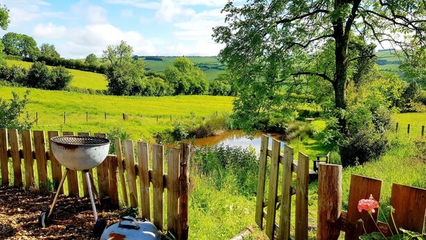 A rustic outdoor setting with a wooden fence, a grill, and a covered pot, overlooking a lush green landscape and a small pond. - Beavers Lodge on top of the Shropshire Hills (Craven Arms)
