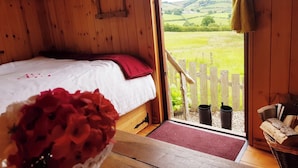 A wooden room with a bed, a window with a view of green fields, and a basket of flowers. - Beavers Lodge on top of the Shropshire Hills (Craven Arms)