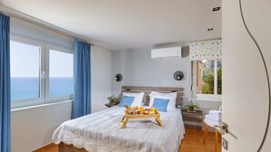 Un dormitorio con una cama grande, una mesita de noche con una bandeja de desayuno, una ventana con vista al mar y un equipo de aire acondicionado.