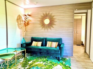 A room with a green sofa, a glass coffee table, a sunburst wall decoration, and a tropical leaf-patterned rug. - Campo Vecchio Sole & Piscine (Ghisonaccia)