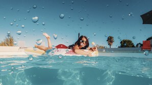A person lying on a float in a swimming pool with clear blue water, surrounded by palm trees and a clear sky. - Campo Vecchio Mare & Piscine (Ghisonaccia)
