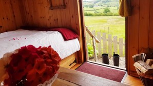 A wooden room with a bed, a window with a view of green fields, and a basket of red flowers. - Cabin Retreat With Beavers on Shropshire Hills (Craven Arms)