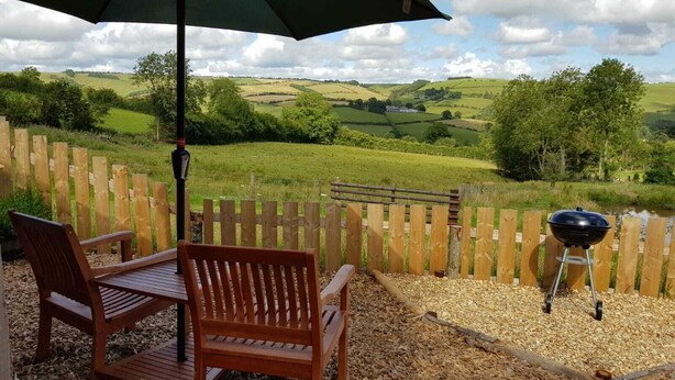 An outdoor seating area with a wooden table and chairs, a barbecue grill, and a view of a green landscape with a wooden fence. - Cabin Retreat With Beavers on Shropshire Hills (Craven Arms)