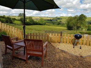 An outdoor seating area with a wooden table and chairs, a barbecue grill, and a view of a green landscape with a wooden fence. - Cabin Retreat With Beavers on Shropshire Hills (Craven Arms)