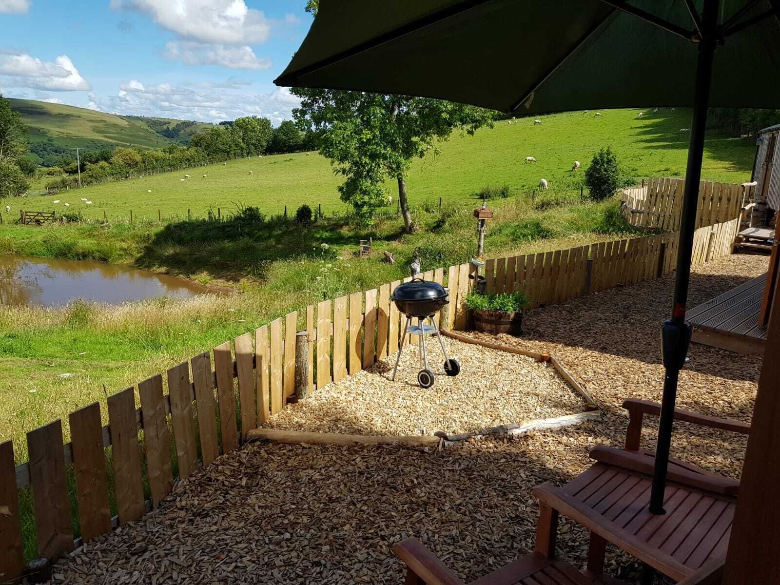 A patio area with a barbecue grill, wooden deck, and a view of a green field with sheep.