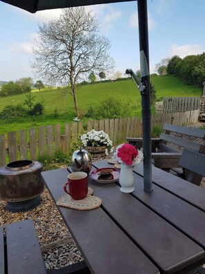 An outdoor seating area with a wooden table, a metal kettle, a red mug, a white vase with pink flowers, and a wooden fence in the background. - Cabin Retreat With Beavers on Shropshire Hills (Craven Arms)
