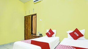 A hotel room with two beds, each with a red and white bedspread and a red pillow with 'oyo' on it. There is a wooden wardrobe and a small table with a water bottle and a box. - OYO 3214 Puri Amalia (Banyuwangi)