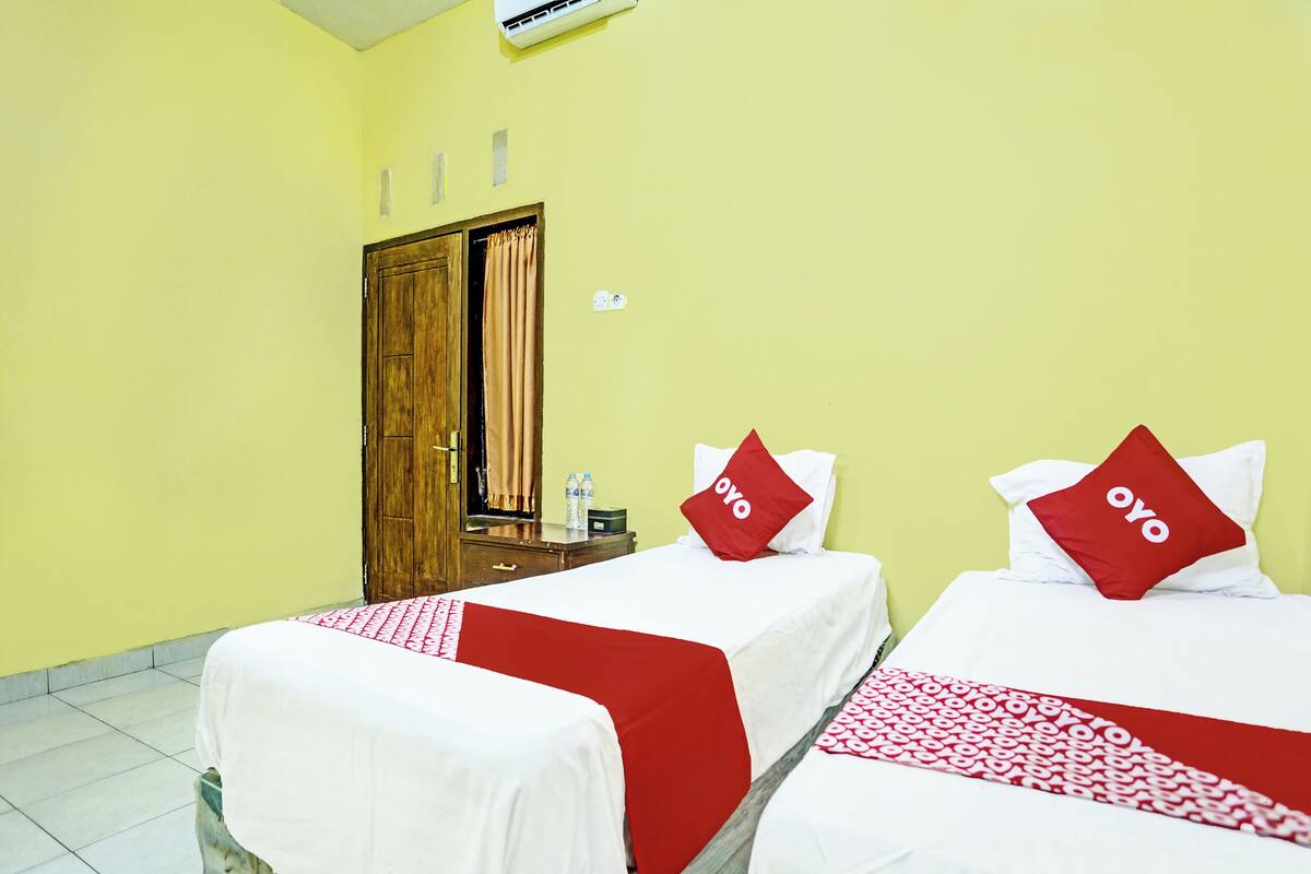 A hotel room with two beds, each with a red and white bedspread and a red pillow with 'oyo' on it. There is a wooden wardrobe and a small table with a water bottle and a box.