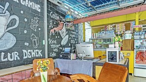 A café with a mural, a coffee machine, a computer, and a table with chairs. - OYO 3214 Puri Amalia (Banyuwangi)
