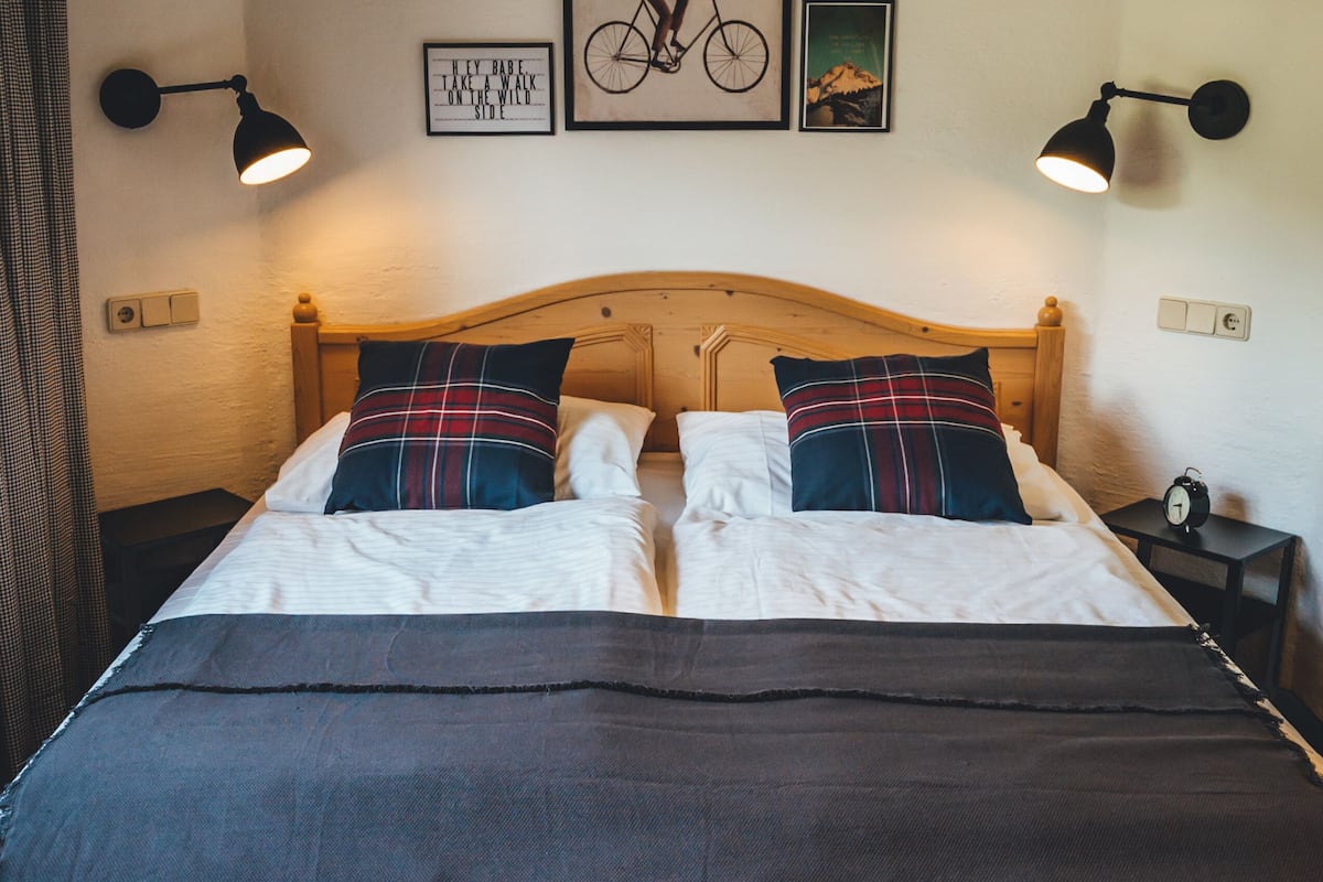 Ein ordentlich bezogenes Bett mit einer karierten Decke, zwei Kissen und einem Holzkopfteil. An der Wand sind Wandleuchten und gerahmte Bilder angebracht.