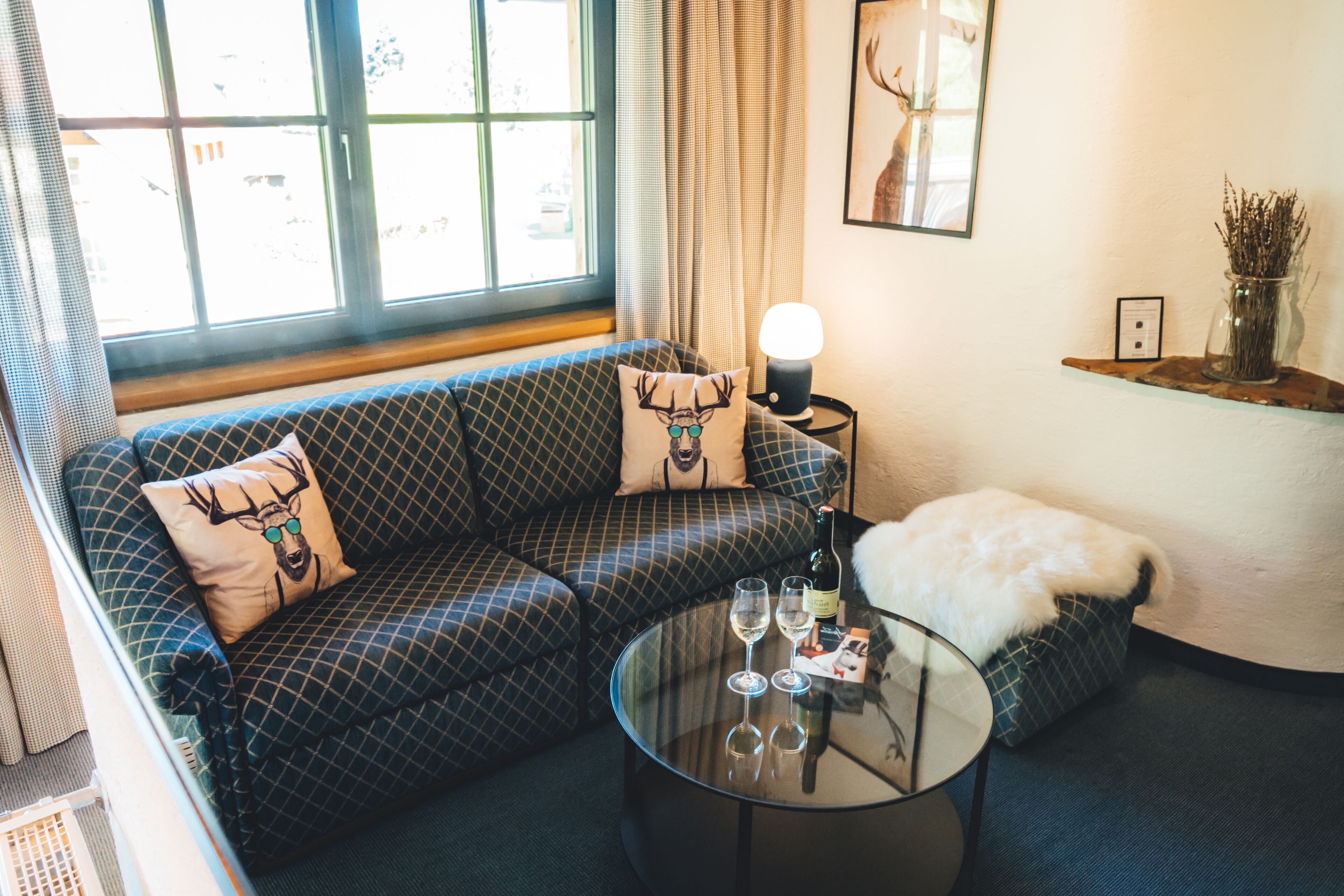 Un salón acogedor con un sofá estampado, una mesa de centro de vidrio con copas de vino, una lámpara y un jarrón decorativo.