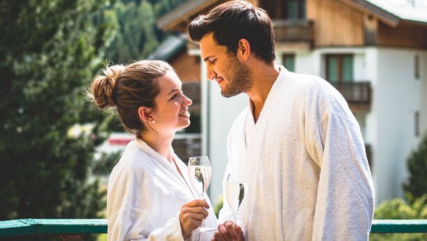 Una pareja con batas blancas de pie en un balcón con montañas al fondo.