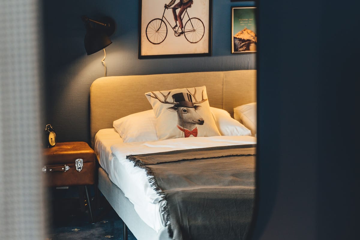 Ein Schlafzimmer mit Bett, Nachttisch, Lampe und Wandbildern, darunter ein Fahrrad und eine Gebirgslandschaft.