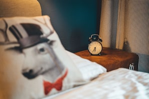 A bed with a pillow featuring a dog illustration, a bedside table with a clock, and a checkered curtain. - Hotel Sportalm (Bad Kleinkirchheim)