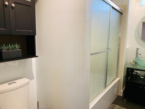 A bathroom with a toilet, a dark cabinet with a plant, a glass shower enclosure, and a sink with a mirror. - The Cottage! Chic Bungalow near Old Town Square! (Fort Collins)