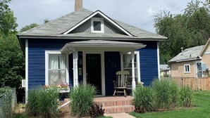 Een huis met twee verdiepingen, met een blauwe gevel, een veranda met een witte stoel, en een meterkast pad dat naar de ingang leidt.