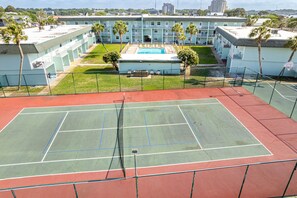 A tennis court with a pool in the background, surrounded by a fence and palm trees. - Venus 314 is a Gulf Side 2 BR on Okaloosa Island by RedAwning (Fort Walton Beach)
