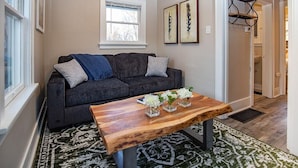 A living room with a dark grey sofa, a wooden coffee table with white flowers, and a patterned rug. - Cozy Carriage House | Walk to UNC | + Free Bikes (Greeley)