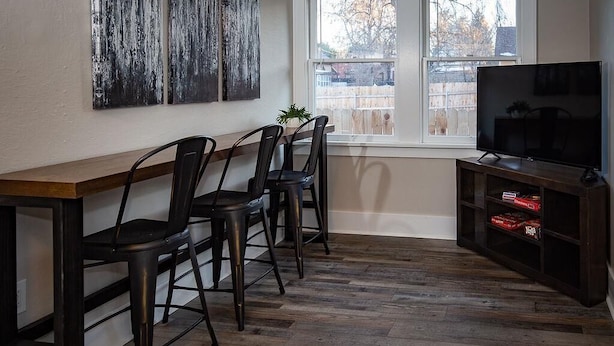 A dining area with a wooden table and chairs, a television on a wooden stand, and a window with a view of a fence. - Cozy Carriage House | Walk to UNC | + Free Bikes (Greeley)