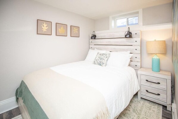 Quarto com cama, mesa de cabeceira, abajur e três quadros de folhas na parede.