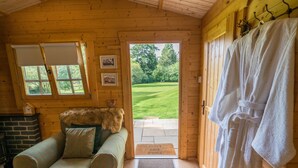 A cozy wooden cabin interior with a green sofa, a fur throw, a fireplace, and a view of a green lawn through a door.