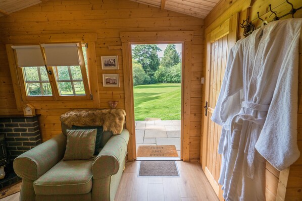 A cozy wooden cabin interior with a green sofa, a fur throw, a fireplace, and a view of a green lawn through a door.