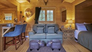 A cozy cabin interior with a wooden ceiling, a plaid sofa, a desk with chairs, and a bed with a green blanket.