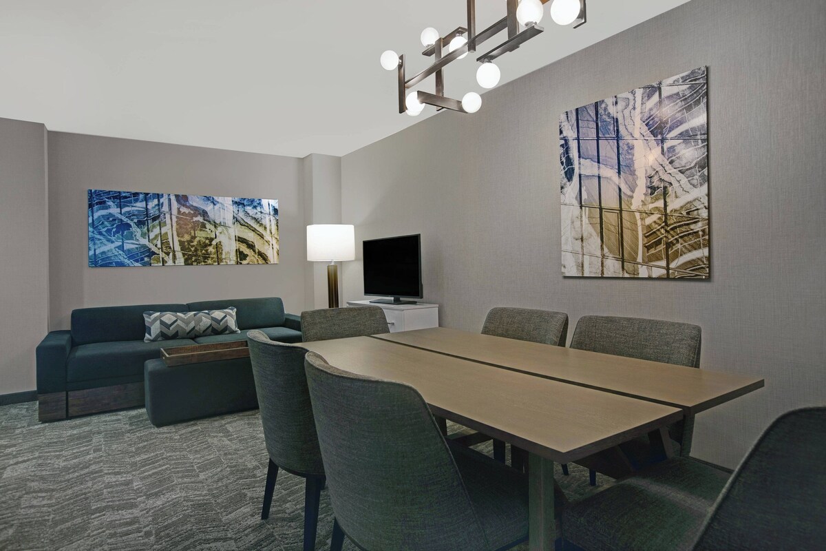A hotel room with a dining table, chairs, a sofa, a TV, and artwork on the walls.