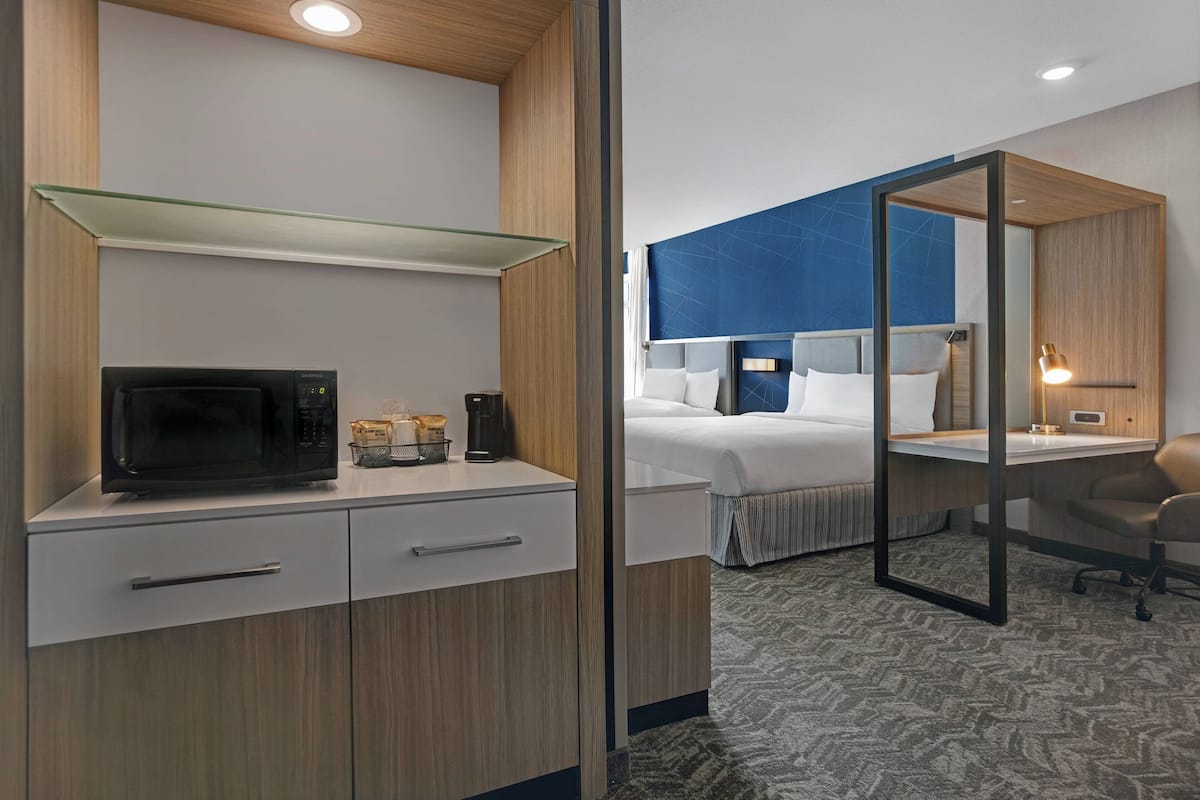 A hotel room with a kitchenette, a bed with a blue accent wall, a desk with a lamp, and a glass partition.
