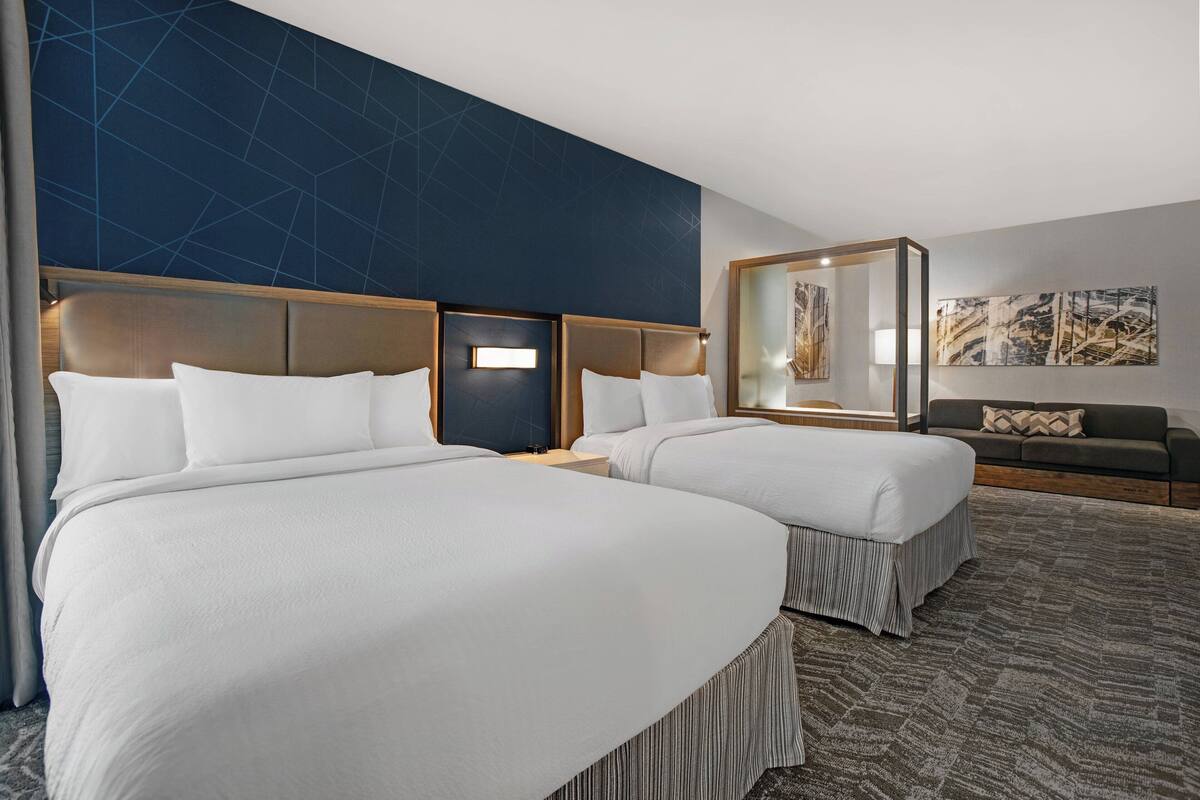 A hotel room with two beds, a dark blue accent wall, a gray sofa, and abstract artwork.