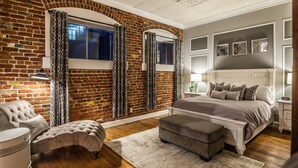 A bedroom with a brick wall, a large bed, a chest of drawers, and a window with curtains. - FireHouse Alley Elegant Loft-Style Retreat (Fort Collins)
