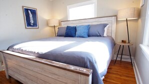 Ein Schlafzimmer mit einem hölzernen Bettgestell, einem großen Bett mit vielen Kissen, zwei Nachttischlampen, einem Fenster und einem an der Wand hängenden Kunstwerk.