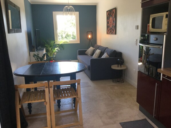 A compact living space with a dining table, chairs, a sofa, a small side table, a potted plant, a hanging light fixture, a clock, and a microwave. - Vacation rental house with pool PUGET LS2-356 (Puget)