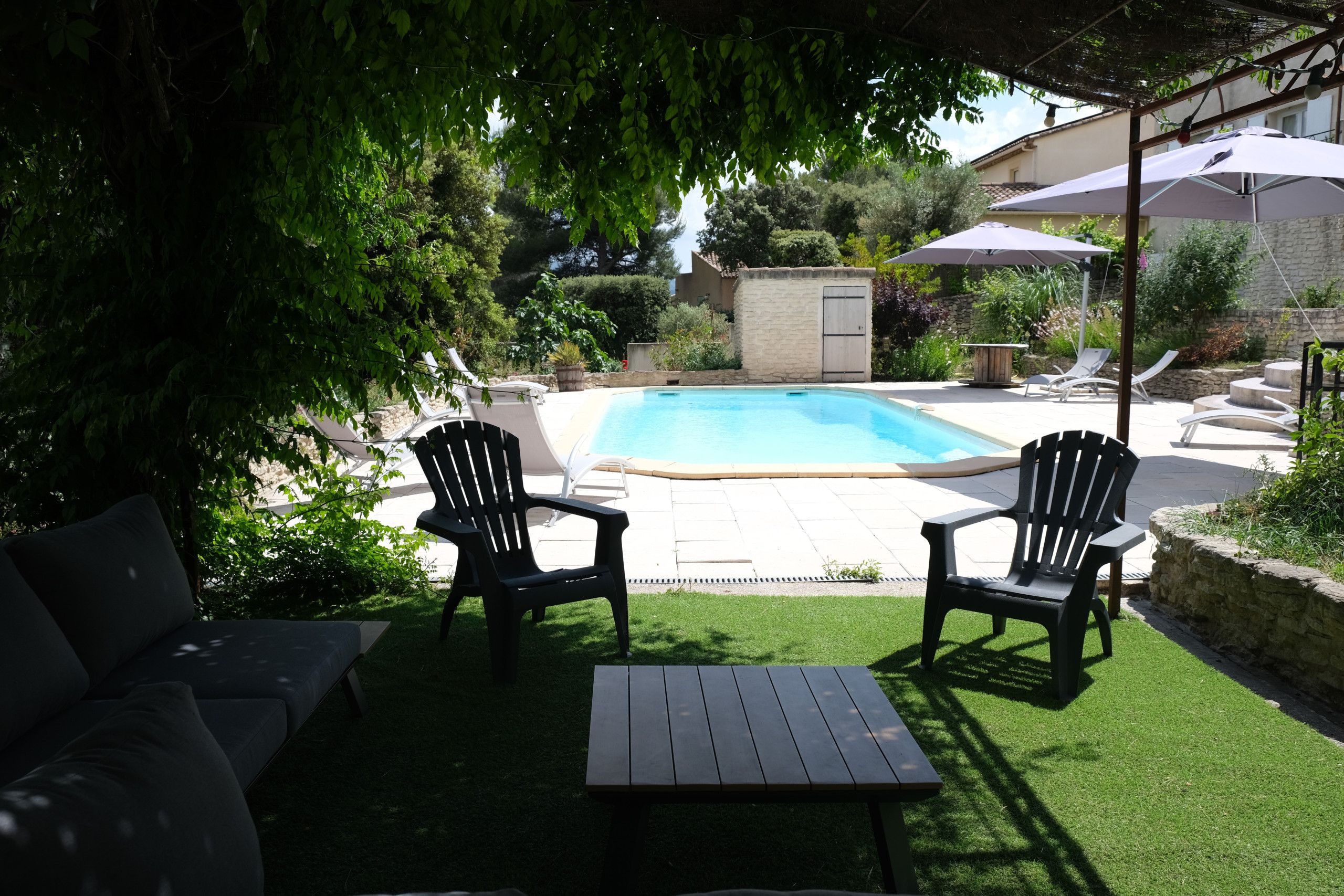 A backyard with a swimming pool, two Adirondack chairs, a wooden table, and a covered patio area.