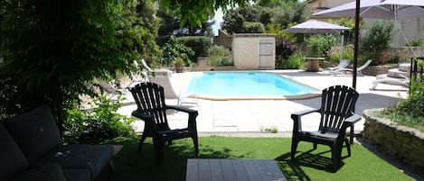 A backyard with a swimming pool, two Adirondack chairs, a wooden table, and a covered patio area.