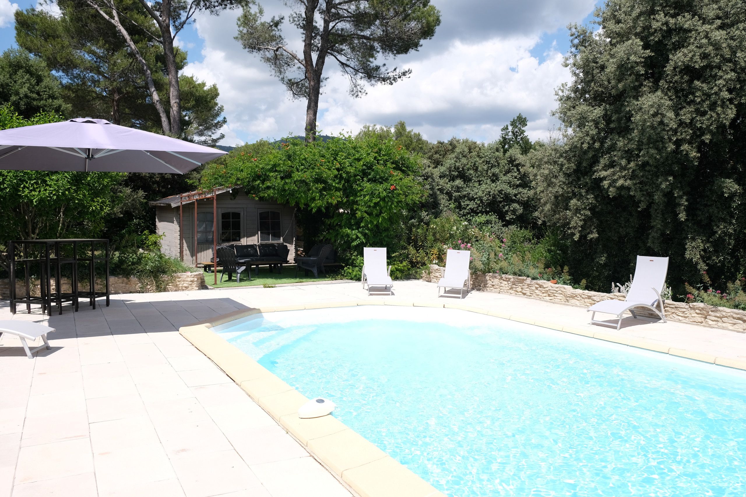 A swimming pool with lounge chairs and an umbrella, surrounded by trees and a stone wall.