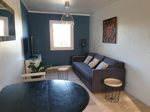 A modern living room with a dark blue wall, a grey sofa with patterned cushions, a round black table, a small wooden stool, and a unique metal wall clock. - Vacation rental house with pool PUGET LS2-356 (Puget)