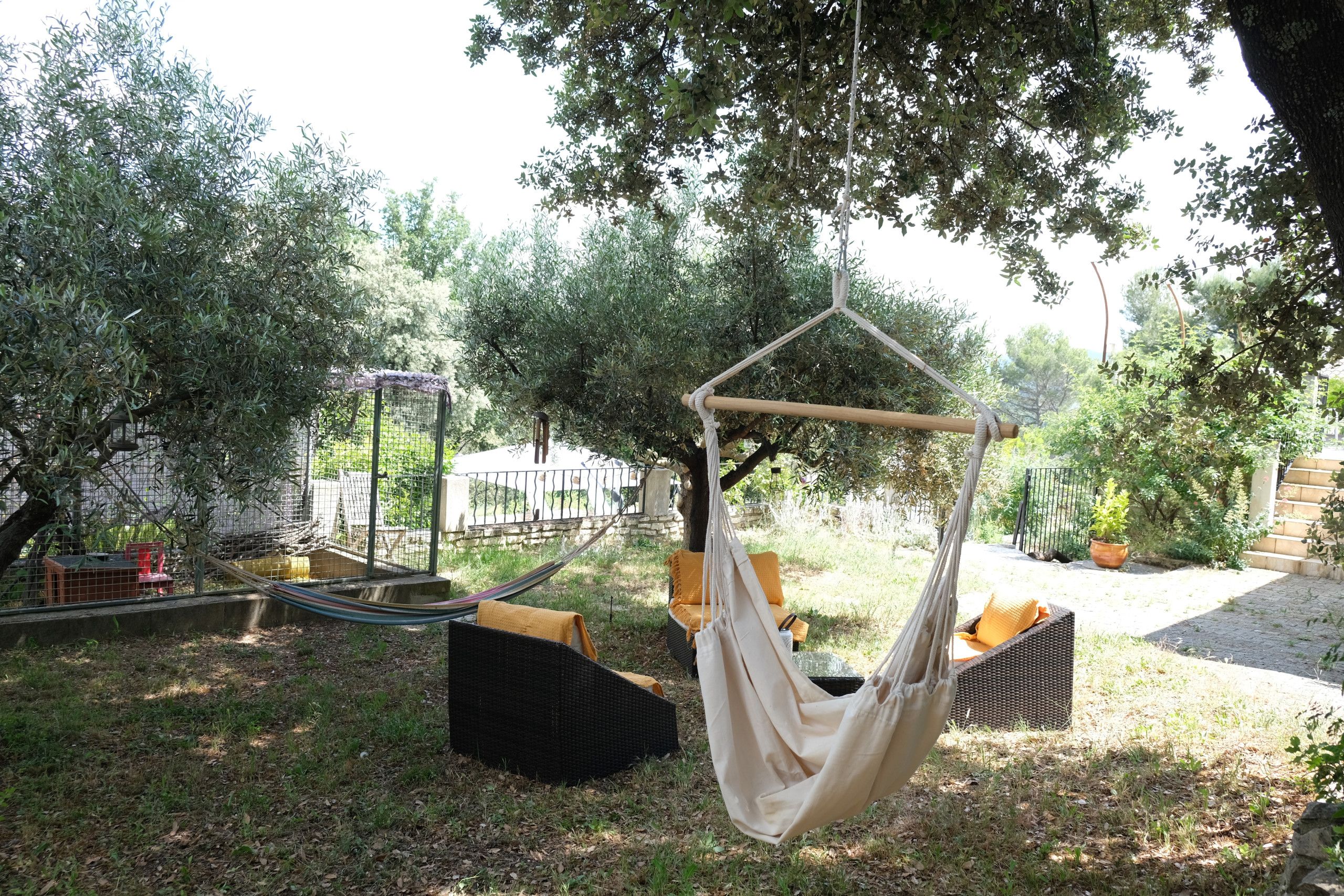 Un fauteuil suspendu, de type hamac, accroché à un arbre, entouré d’un jardin clos, et un fauteuil en osier avec des coussins.