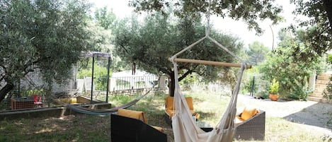 Una silla hamaca colgada de un árbol, rodeada por un jardín con una cerca y una silla de mimbre con cojines.