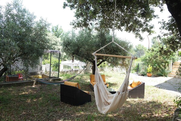 A hammock chair suspended from a tree, surrounded by a garden with a fence, and a wicker chair with cushions. - Vacation rental house with pool PUGET LS2-356 (Puget)